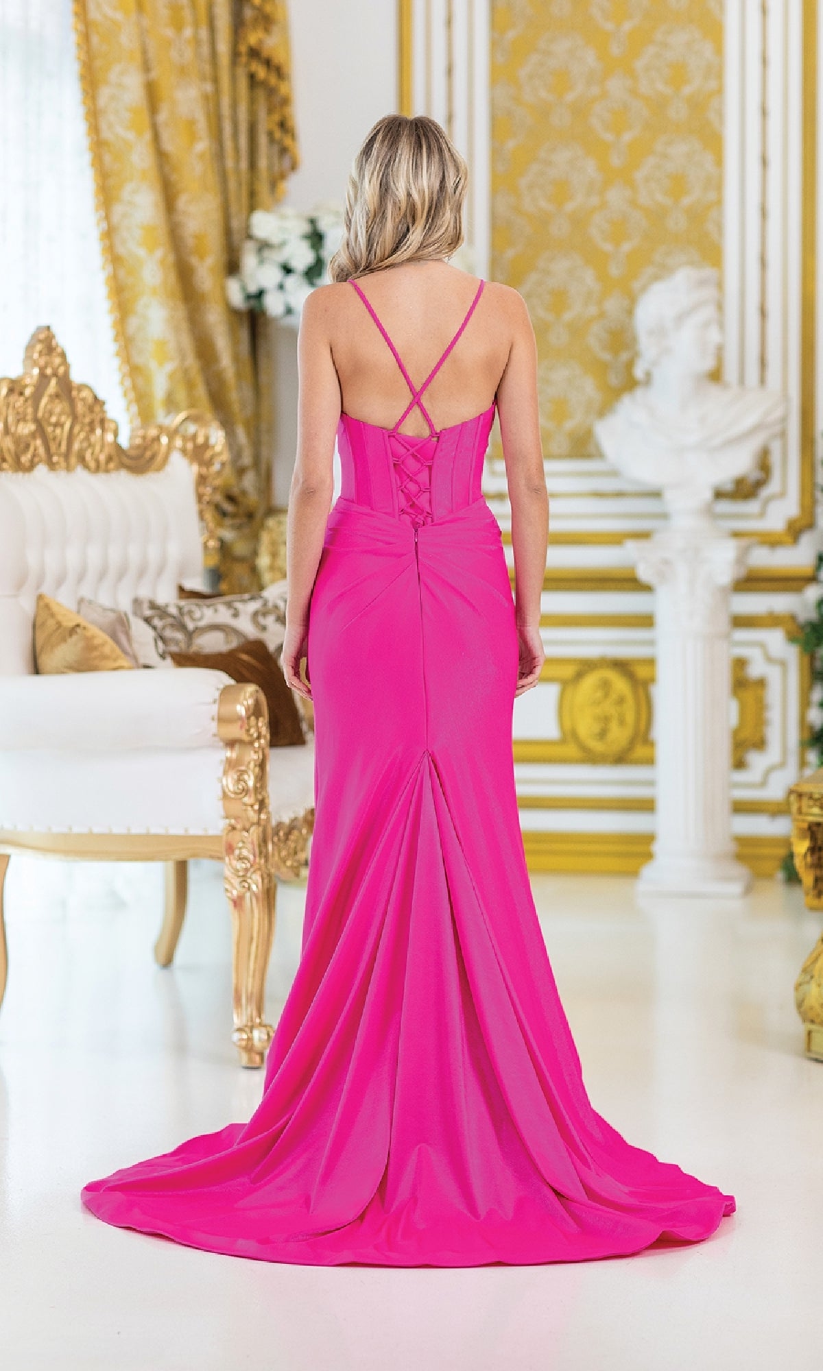 A woman in a Lace-Up Long Corset Prom Dress by Dancing Queen 4456, bright pink and elegant, stands with her back to the camera in an ornate room with gold accents, white furniture, and classical decor.